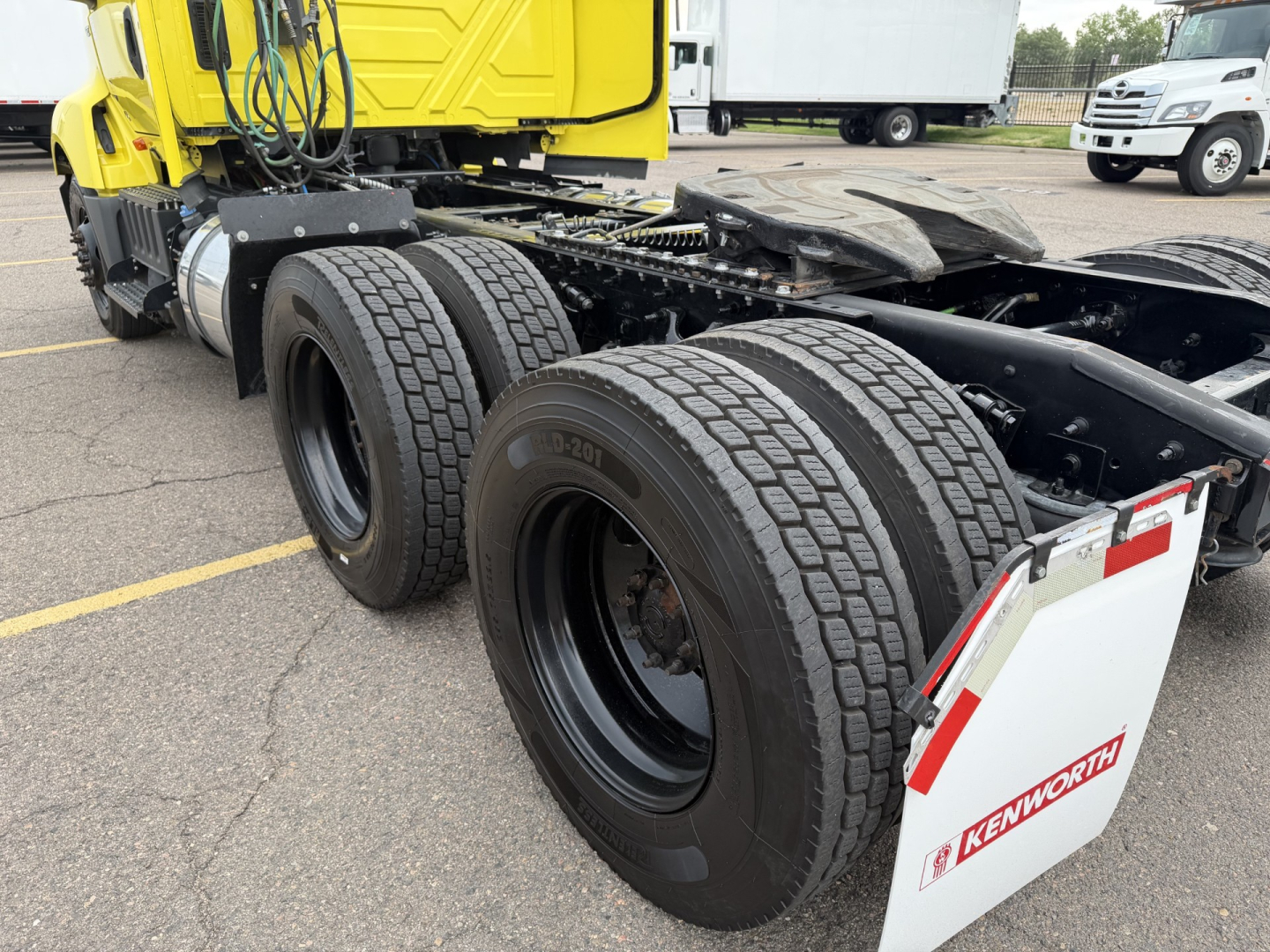 Rear view of a 2023 International LT625 Conventional Daycab truck showing dual rear axles with large black tires and Kenworth mud flaps; yellow cab in background.