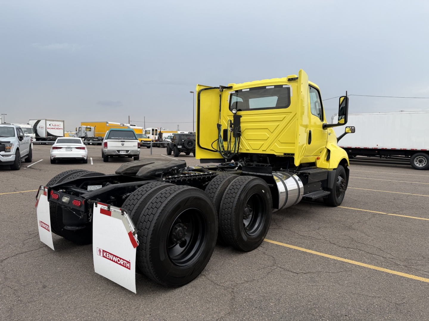 Truck tractor rear view showing dual rear axles and fuel tanks with bright yellow cab in a parking lot.
