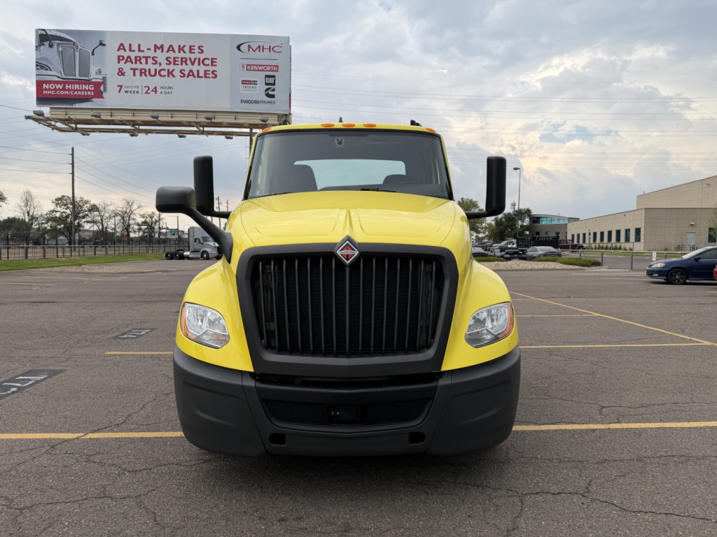 Front view of a yellow 2023 International LT625 conventional daycab truck with black grille, large bumper, side mirrors, and dealership lot in background.