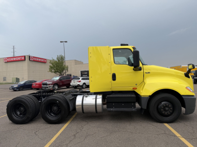 Truck chassis with a bright yellow cab and black undercarriage. Side view showing fuel tanks, dual rear axles, and large tires. Background shows a Kenworth dealership.