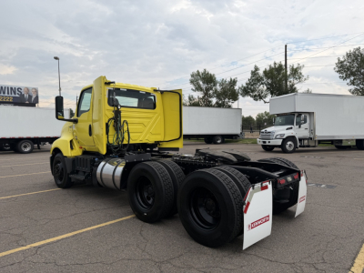 Truck chassis cab in a parking lot with long sleeper area missing; bright yellow International LT625 front cab, dual rear axles, white mud flaps, fuel tanks, and surrounding trucks.
