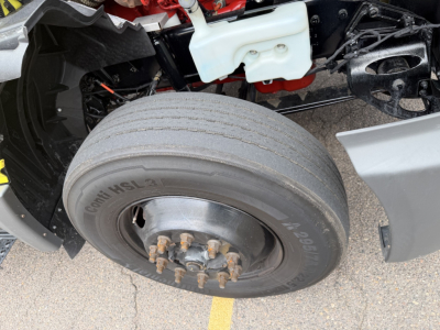 Wheel and front axle area of a used 2023 International LT625 conventional daycab showing a single front tire and hub with lug nuts; partial engine bay components and coolant reservoir visible.