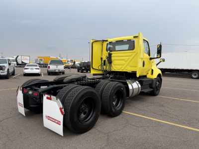 Truck tractor rear view showing dual rear axles and fuel tanks with bright yellow cab in a parking lot.
