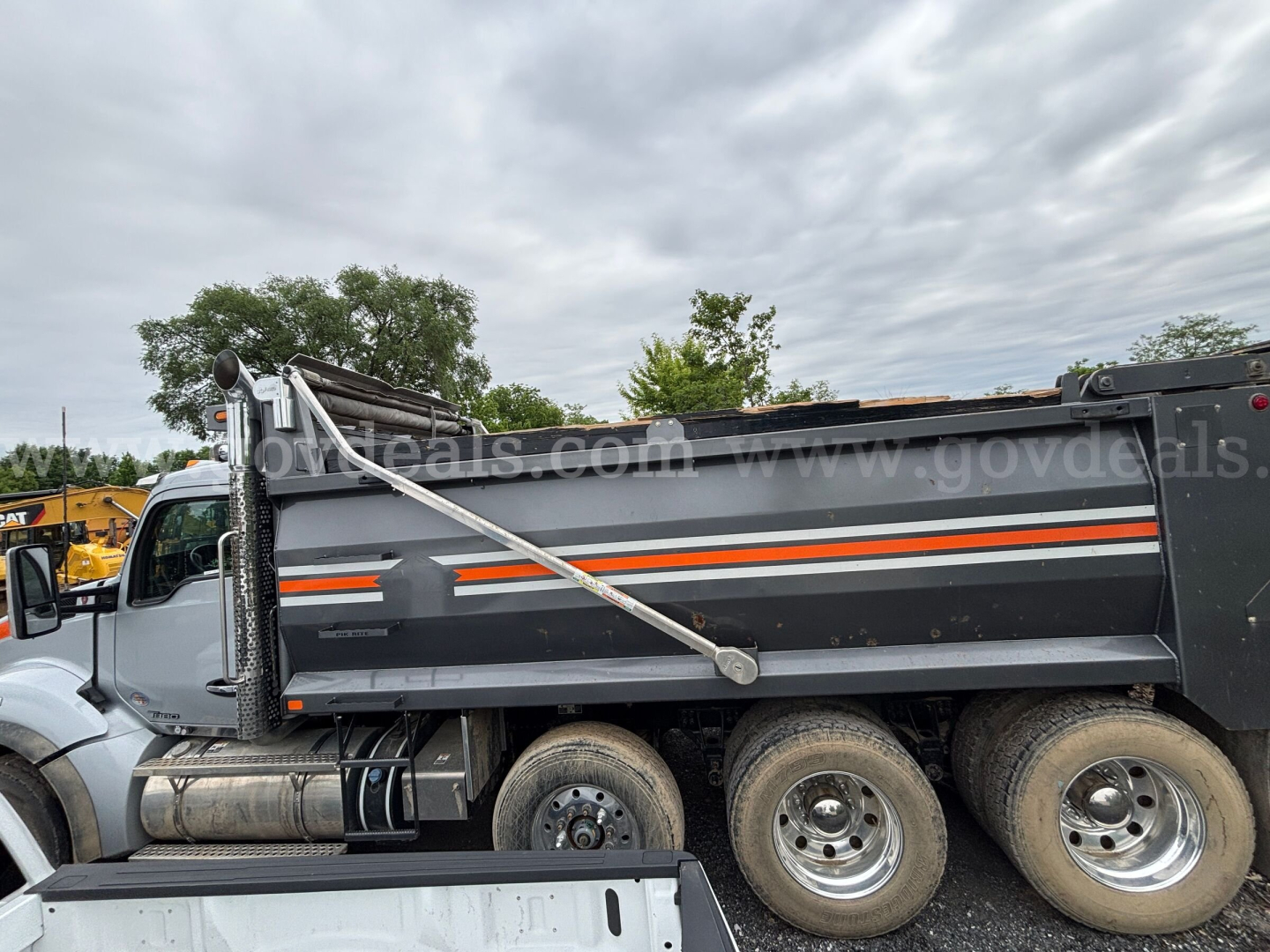 Dump truck side view showing International 7400 dump bed with orange stripe, dual rear wheels, and hydraulic lift cylinder.