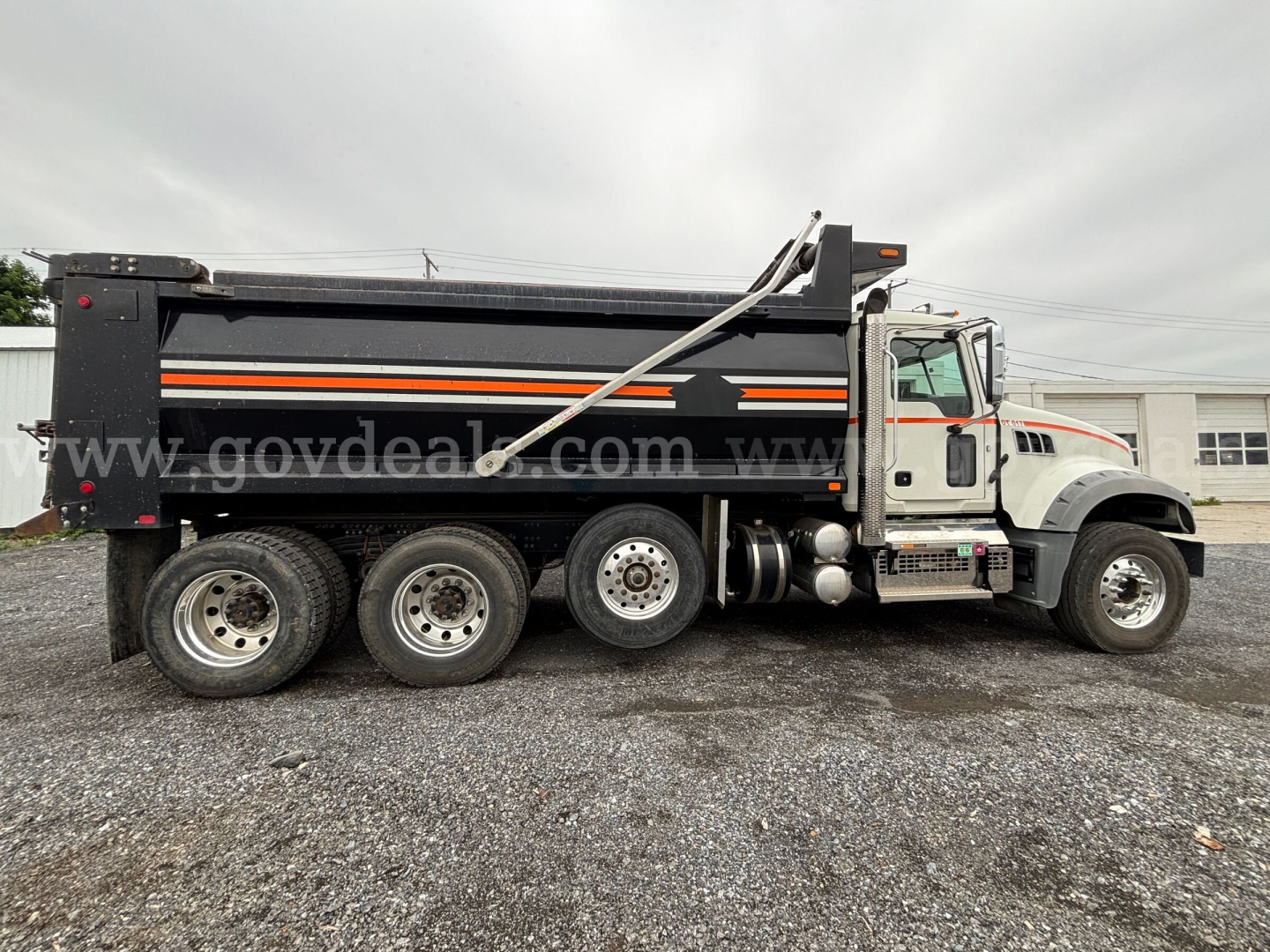 Dump truck side view showing International 7400 heavy-duty chassis with black dump body and orange stripes, six-wheel configuration, and hydraulic lift on a gravel lot.