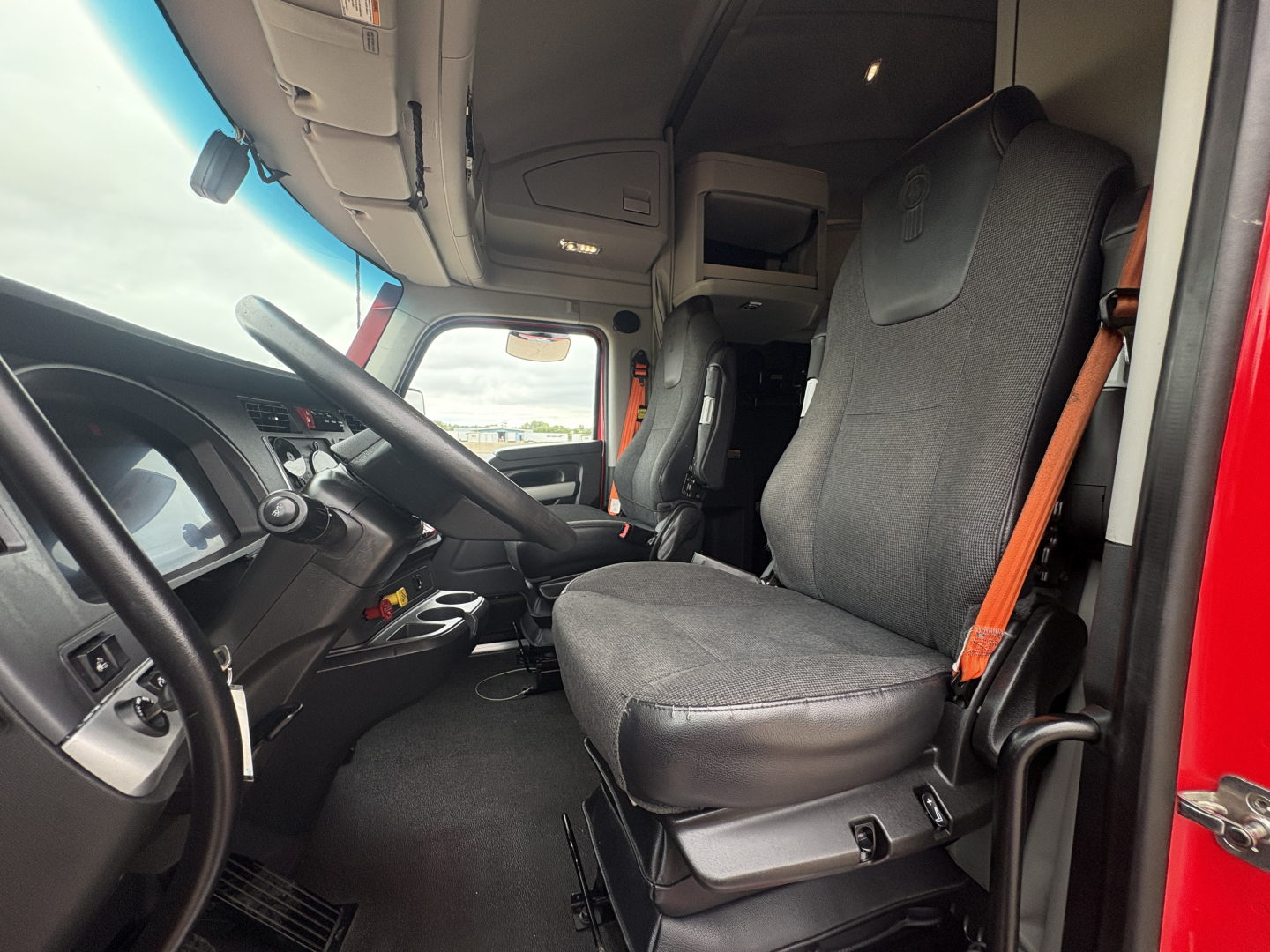 Kenworth T680 cab interior showing driver seat and dashboard with steering wheel and exposed overhead storage.