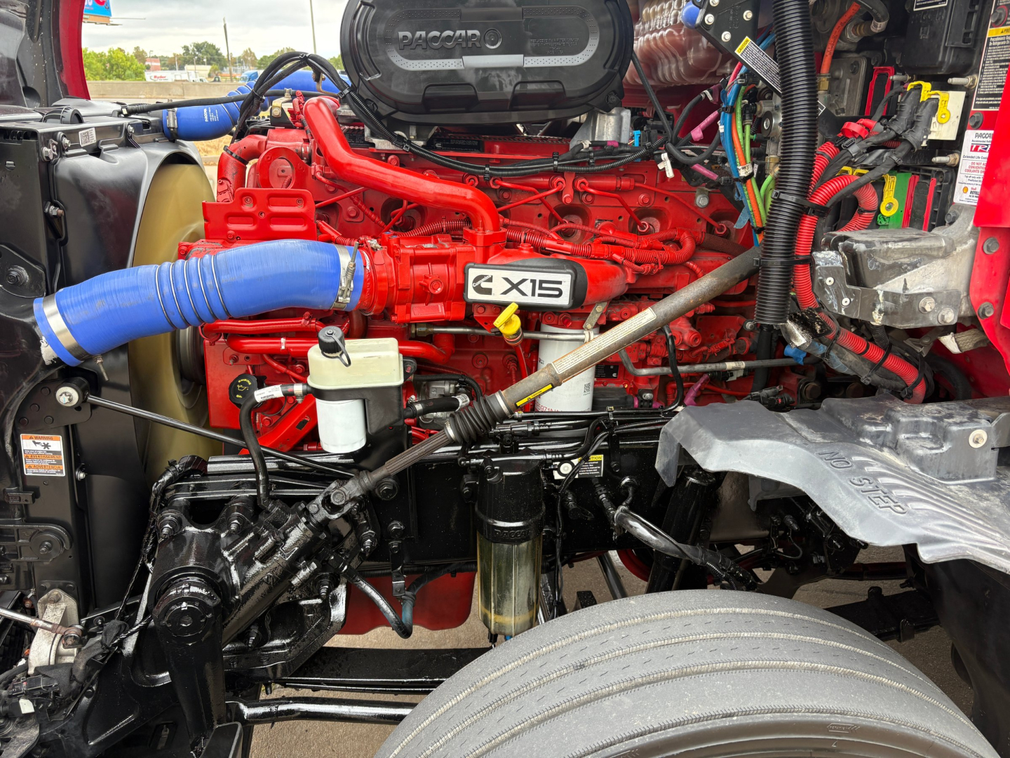 Kenworth T680 engine bay with bright red engine, blue intake hose, and various hoses and wiring visible under hood.