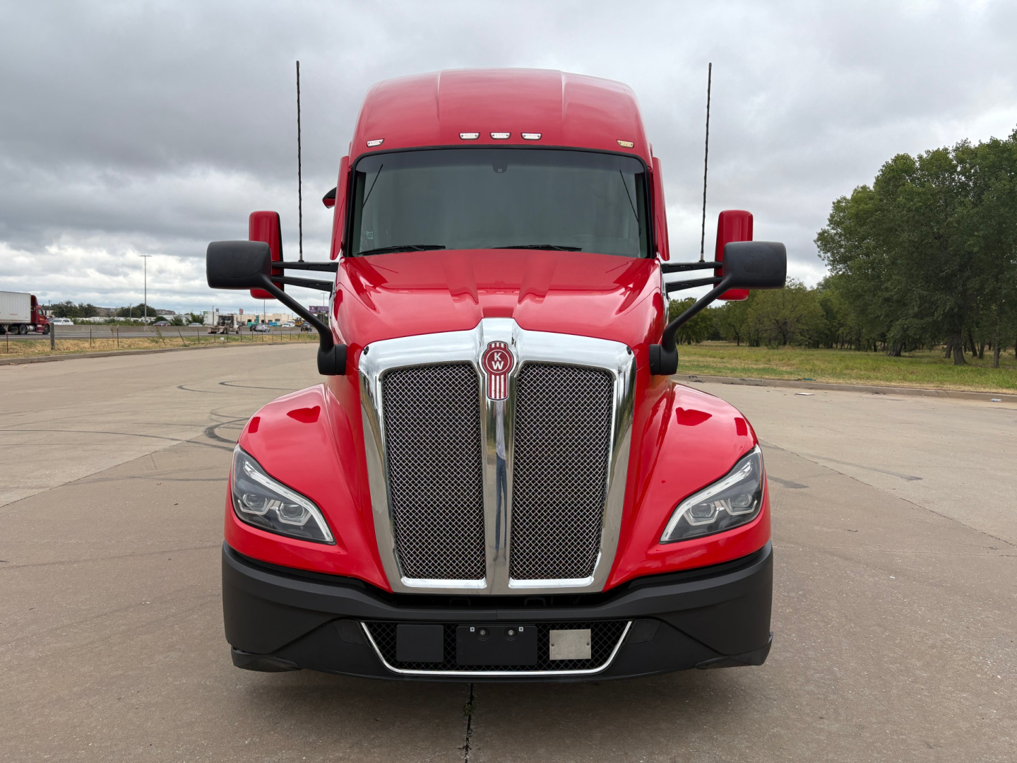 Kenworth T680 front view showing bold chrome grille, large headlights, red cab, black bumper, and side mirrors.