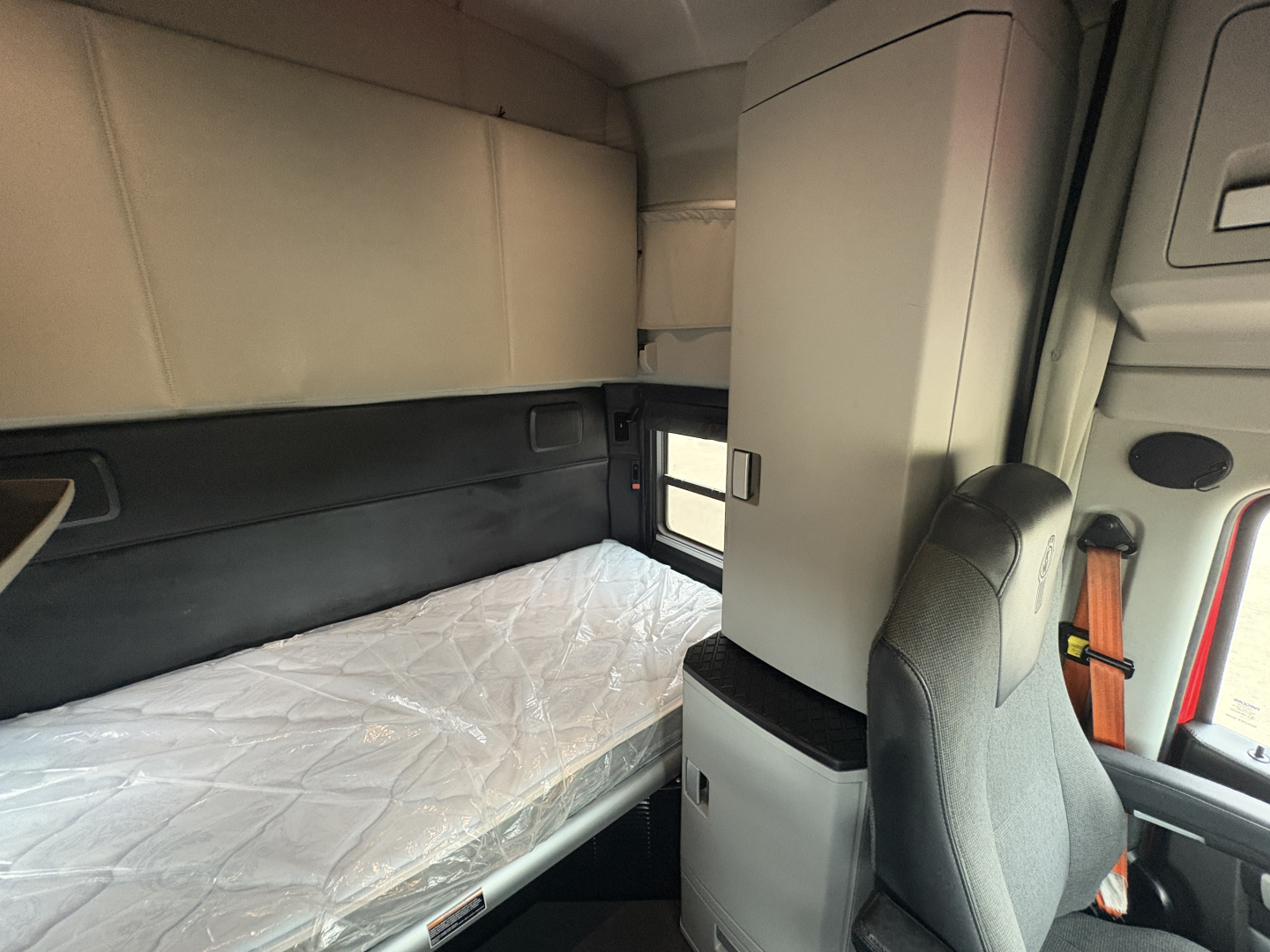 Truck cab interior with sleeper bunk visible, single bed with plastic-wrapped mattress, cabinet, window, and passenger seat in view.