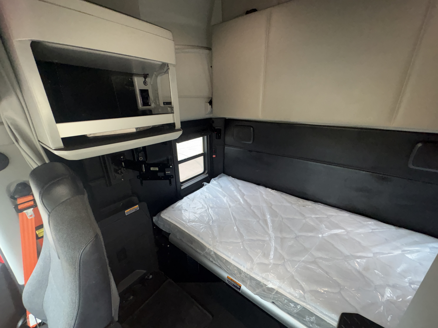 Kenworth T680 sleeper cab interior showing a single bunk bed and overhead storage unit with a small window and seat/console area.