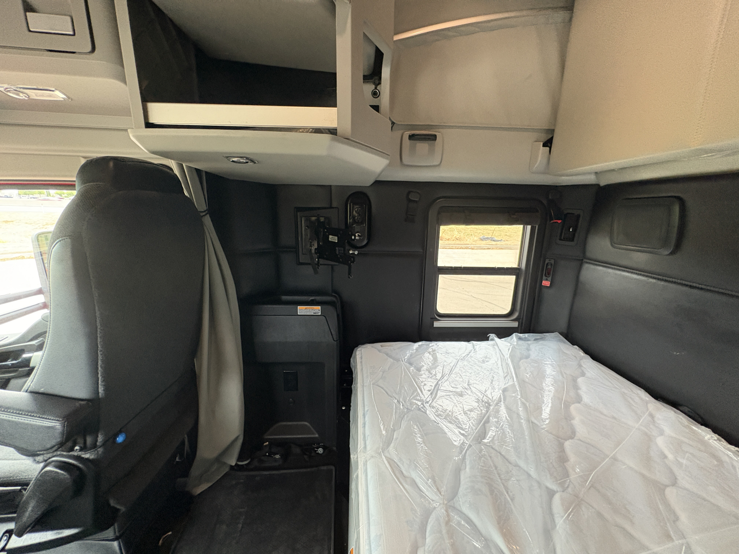 Kenworth T680 sleeper cab interior showing bunk with plastic cover, overhead storage, and window.