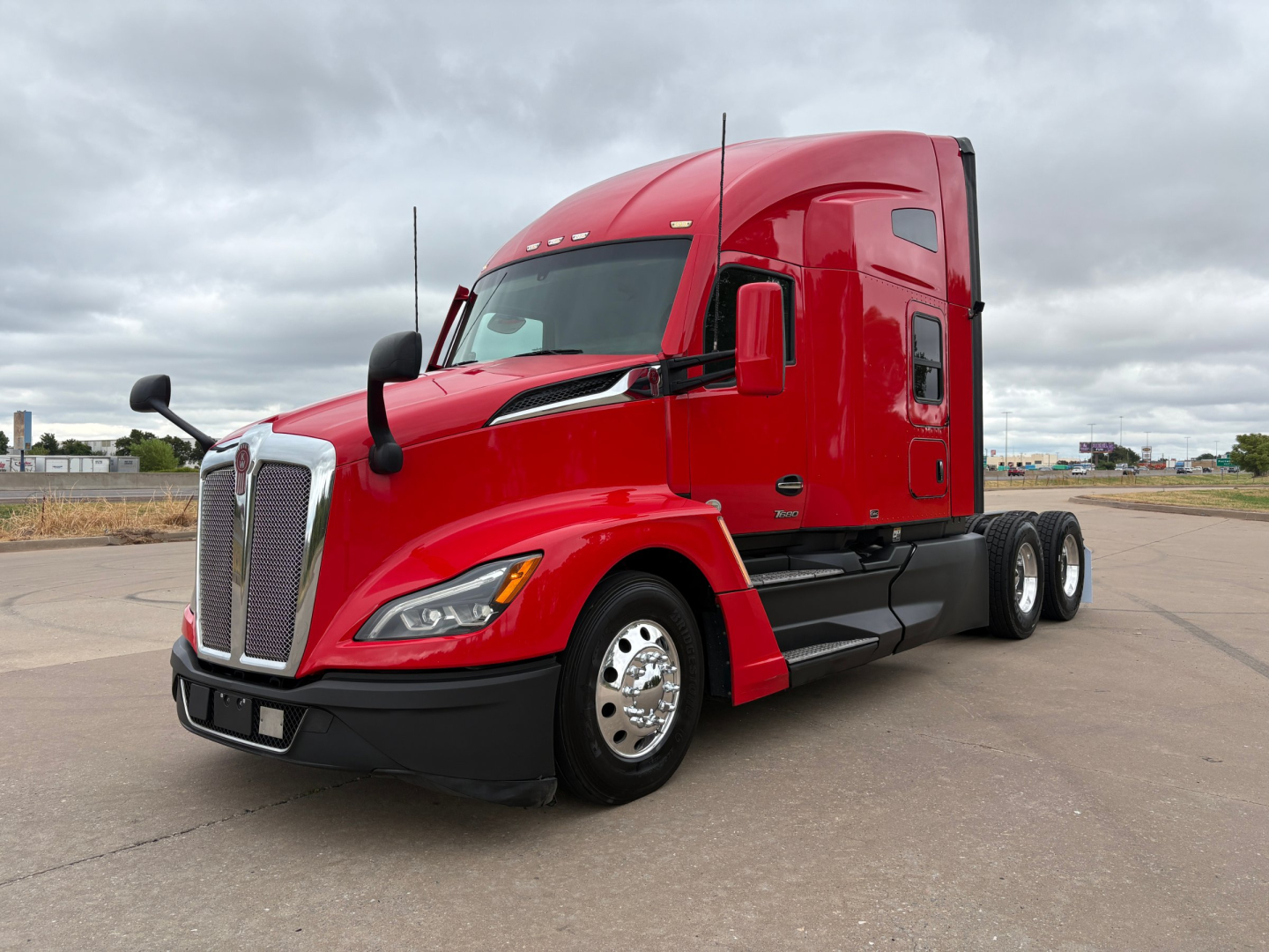 Red Kenworth T680 tractor with sleeper, side view showing front grille, headlights, large mirrors, and dual rear axles.