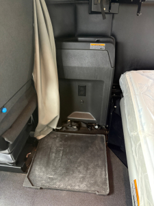 Kenworth T680 truck interior shows a compact appliance/console with fold-out step and storage area; factory label on black console; light gray fabric seat to the right; beige curtain left.