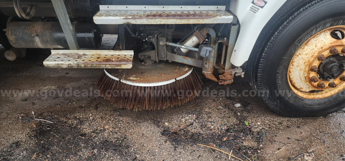 Sweeper truck underside showing rotating cylindrical brush and side steps with rusted metal frame.