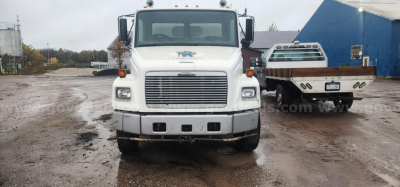 Front view of a white Freightliner FL70 truck with two orange marker lights on hood, large grille, bumper, and two side mirrors; another utility truck behind; overcast lot.