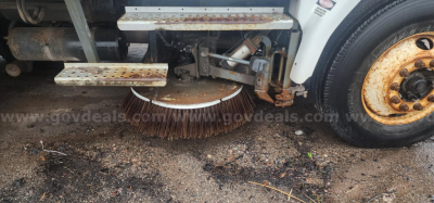 Sweeper truck underside showing rotating cylindrical brush and side steps with rusted metal frame.