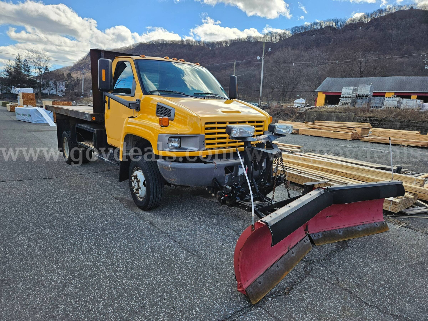 2005 GMC C4500 flatbed dump with yellow cab and red snow plow attachment in a lumber yard.
