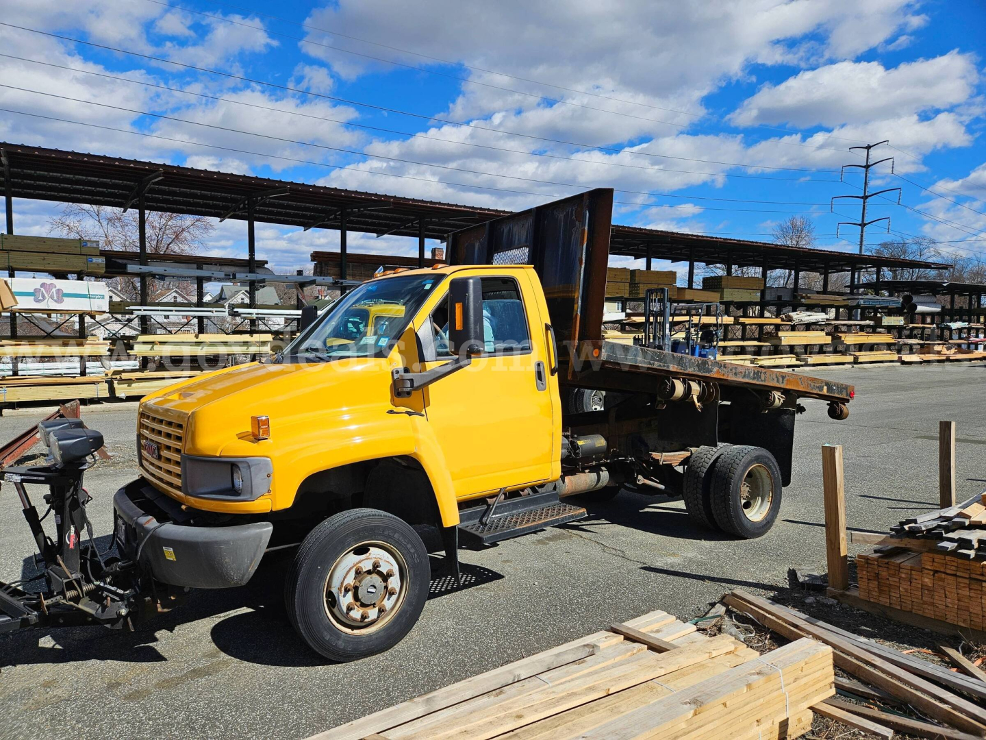 2005 GMC C4500 flatbed dump truck in yellow with a raised bed, cargo yard with wood planks in foreground.