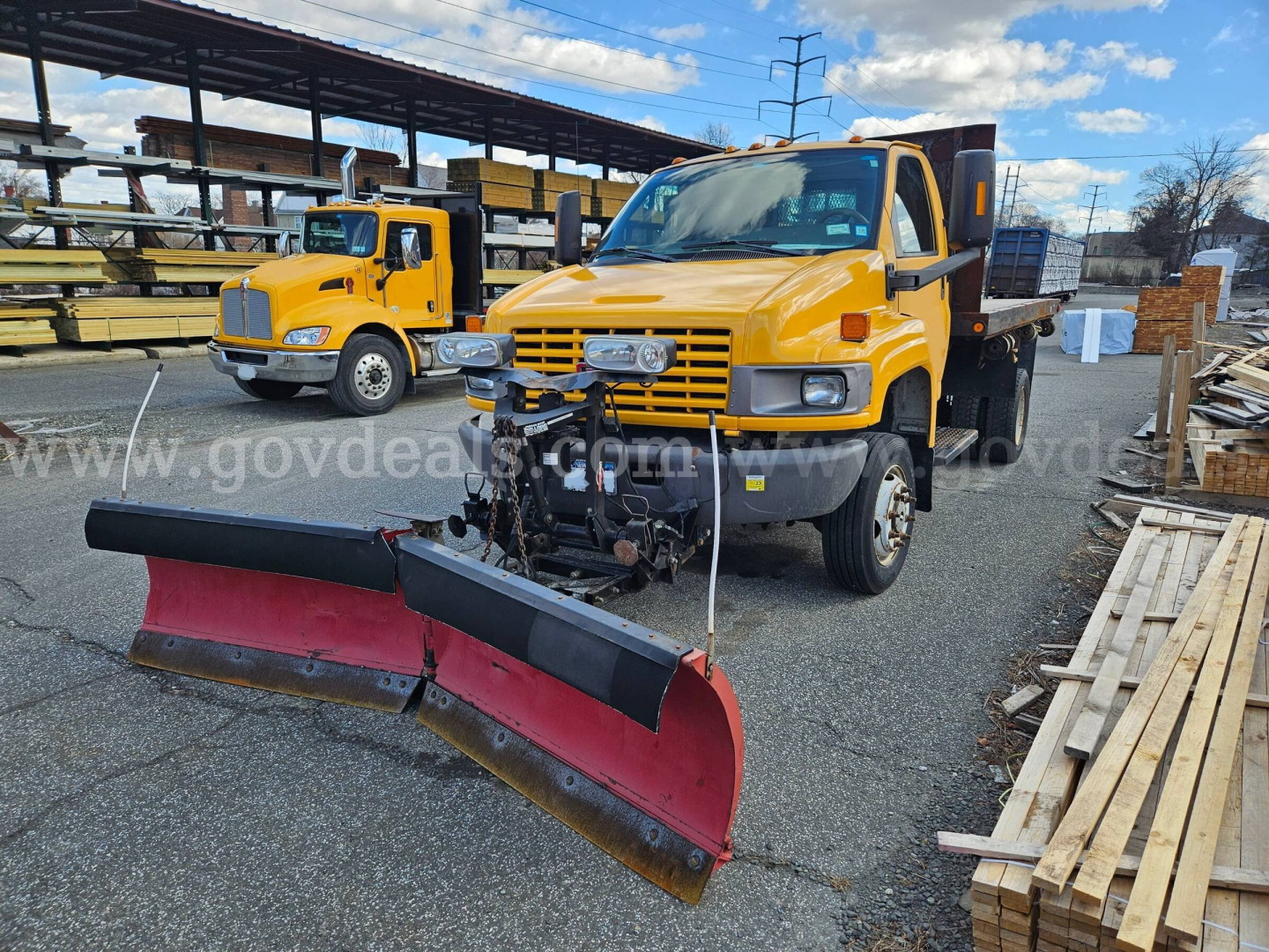 GMC C4500 flatbed dump truck with front mounted plow and yellow cab, outdoor yard with wood planks in background.