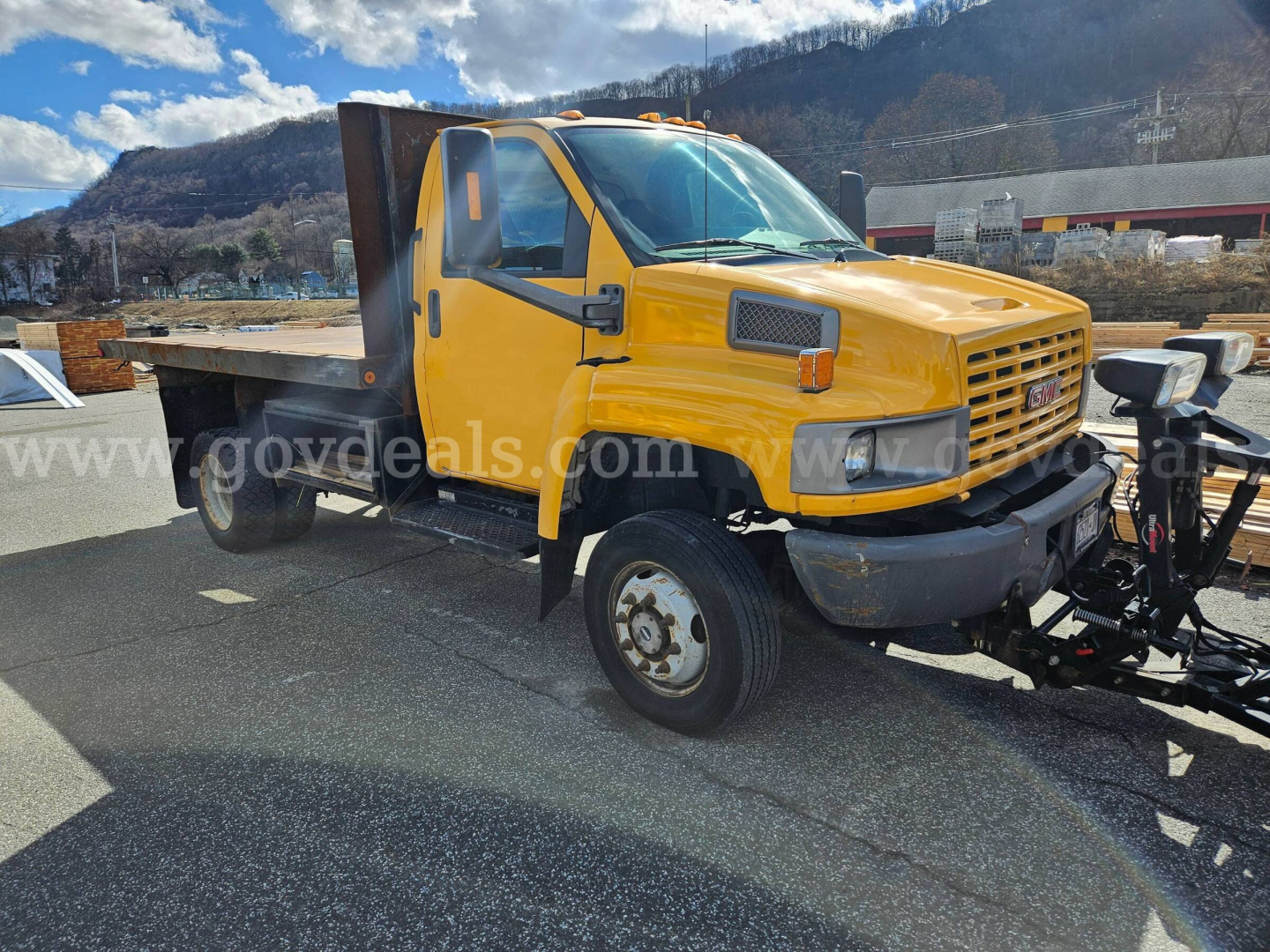 GMC C4500 flatbed dump truck in yellow, front and side view showing cab, dual rear wheels, and equipment on flatbed.