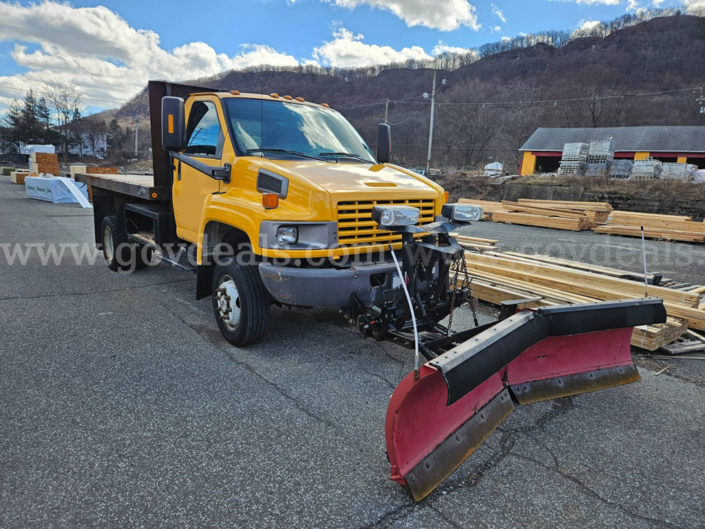 2005 GMC C4500 flatbed dump with yellow cab and red snow plow attachment in a lumber yard.