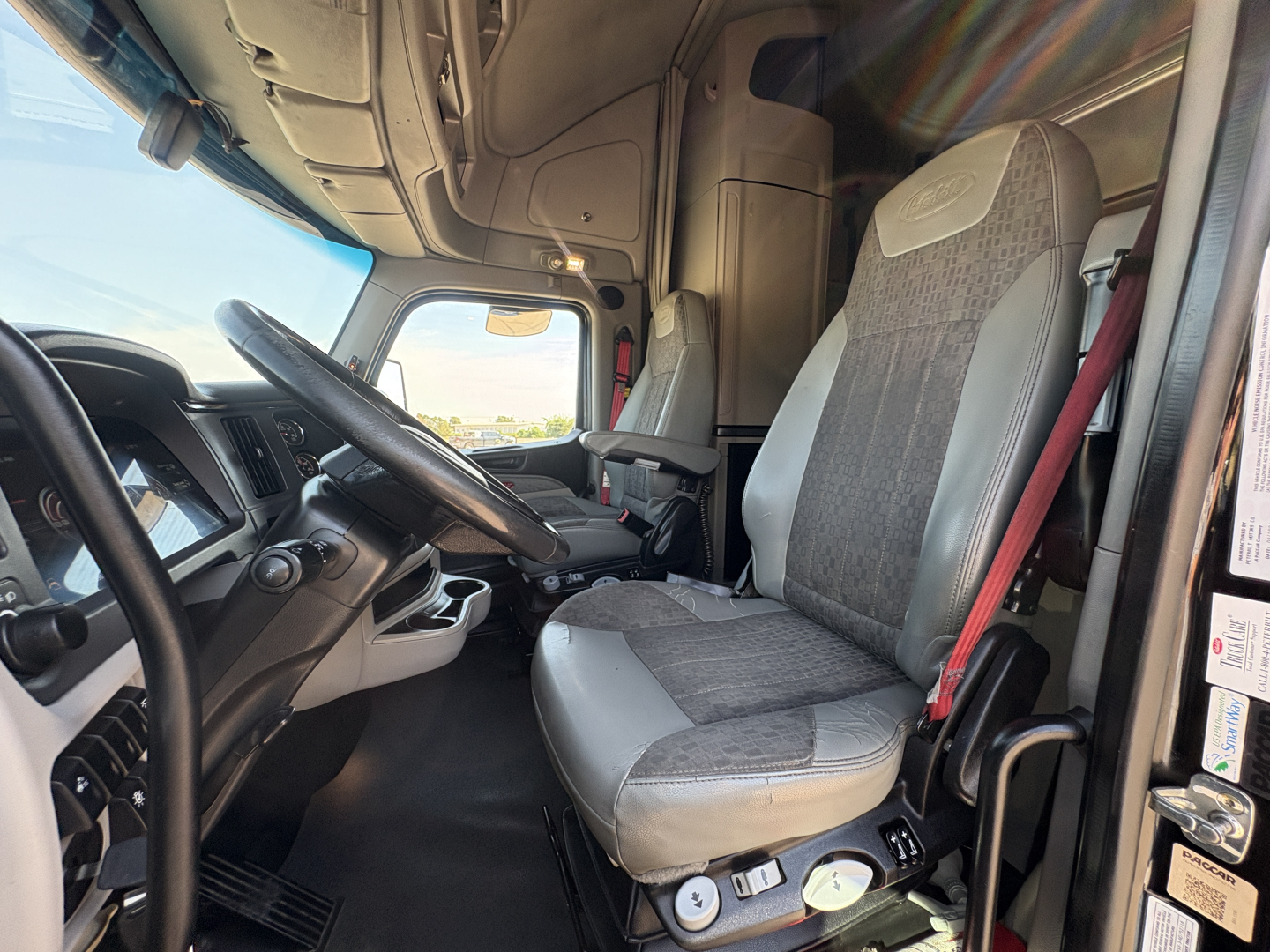 Truck cabin interior of a 2022 Peterbilt 579, driver seat and steering wheel visible, dashboard and overhead panel, light gray fabric seat with textured inserts.