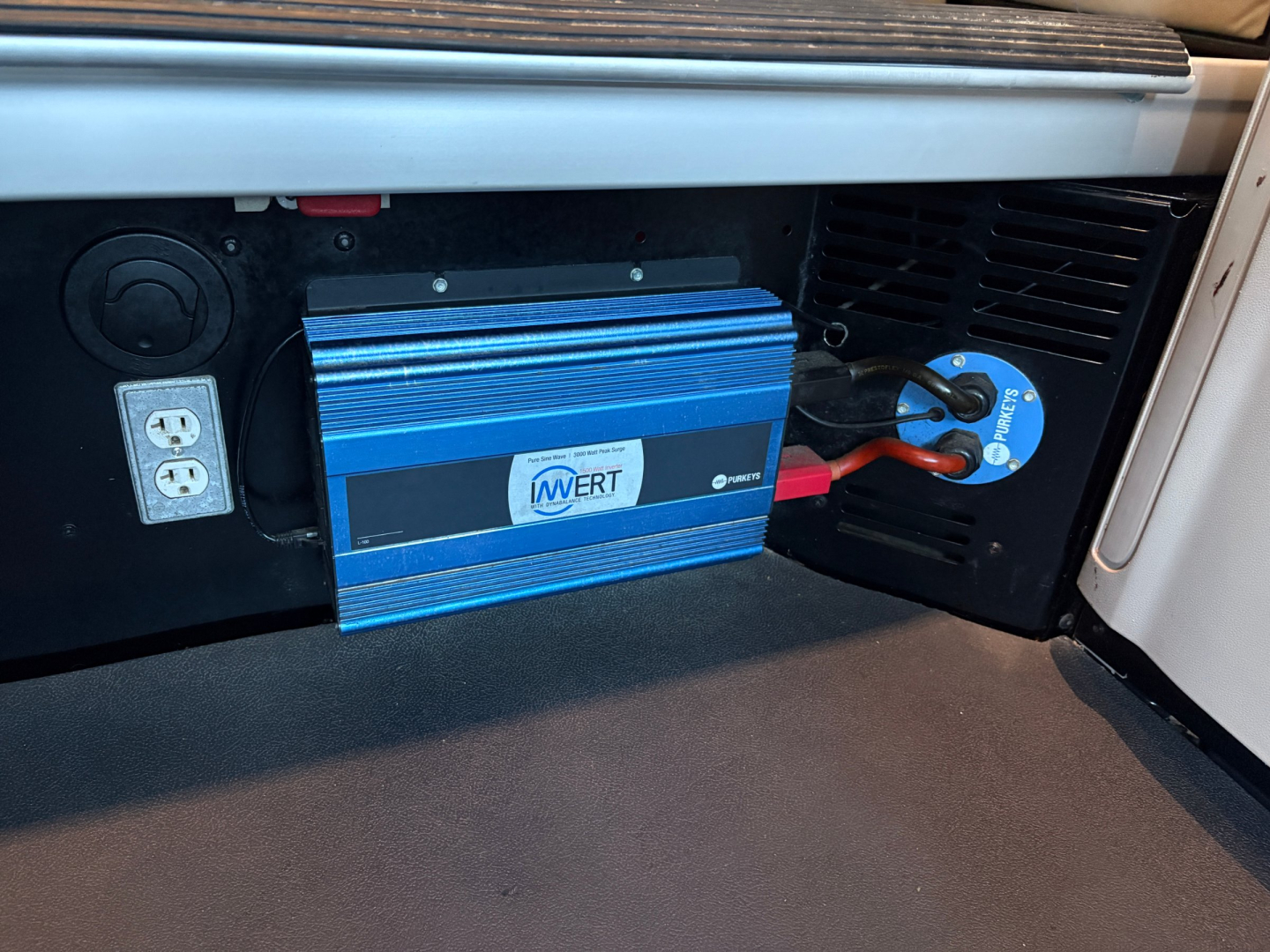 In-cab area of a Peterbilt 579 showing a blue portable inverter/charge device mounted under the dash with a circular cigarette lighter style outlet and a blue ringed plug connected to a power receptacle.