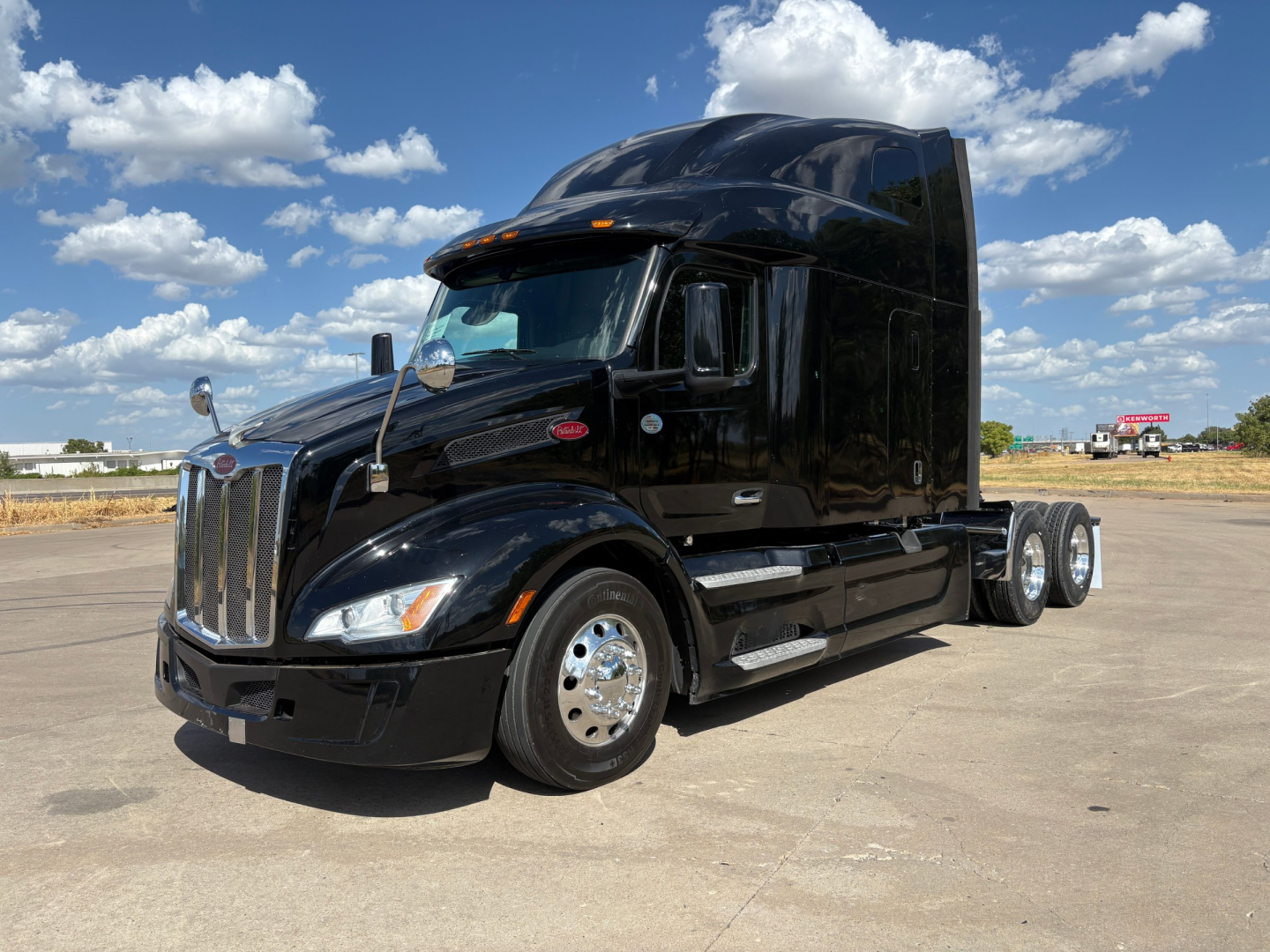 2022 Peterbilt 579 semi-truck in black, cab-over-high-roof sleeper with chrome wheels, parked on pavement under blue sky with clouds; side view shows dual rear axles and polished fuel tanks.