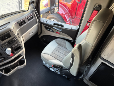 Peterbilt 579 truck cab interior showing driver seat and dashboard with controls, steering wheel, and window view; part of exterior red truck visible through window.