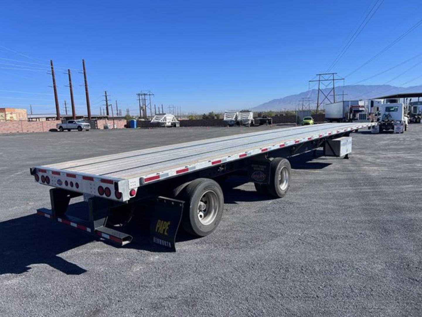 Flatbed trailer with dual rear axles in a parking lot, long aluminum deck, white and red reflective tape along edges, undercarriage and mud flaps visible.