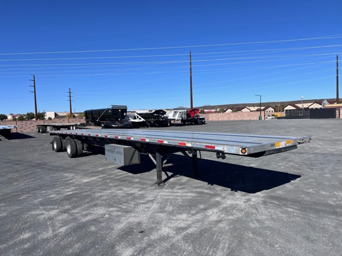 Flatbed trailer in a parking lot showing a long aluminum deck with dual rear axles and support legs.
