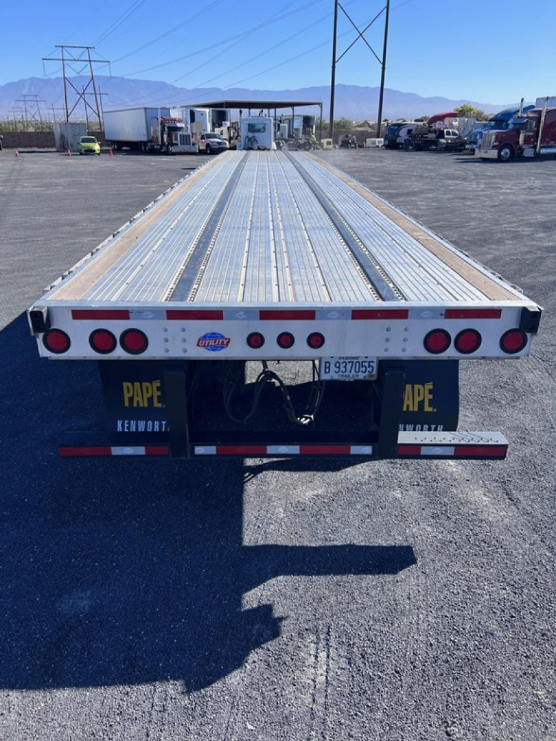 Flatbed trailer showing long aluminum deck with perforated surface and rear bumper with multiple lights and red/white reflective tape.
