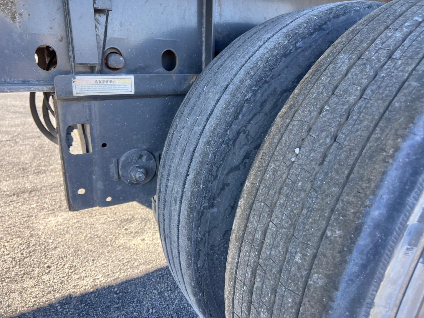 Trailer wheel and frame close-up showing dual rear axles with thick tread tires and metal bracket with warning label.