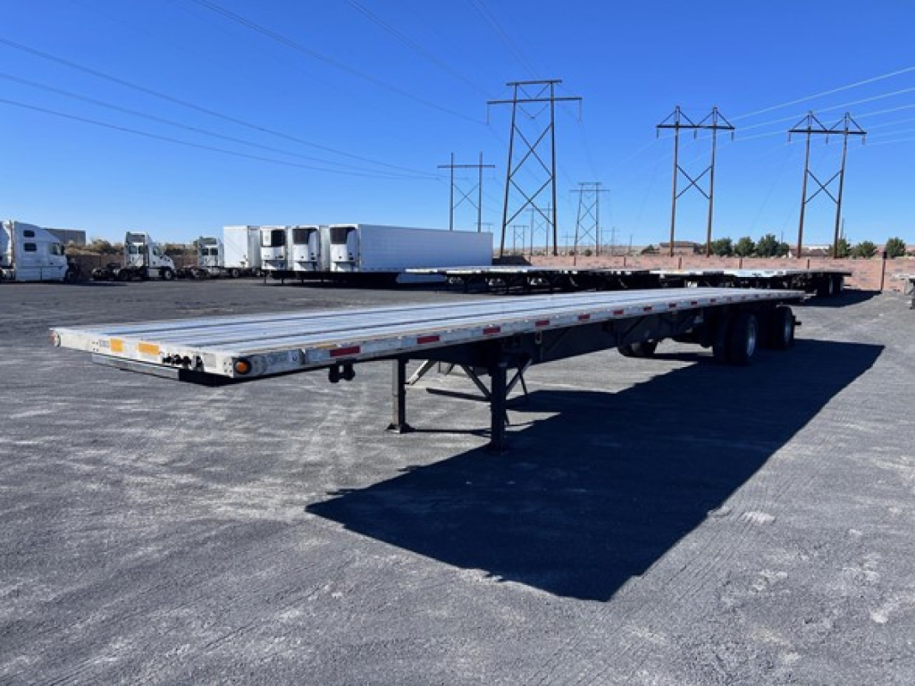 Utility flatbed trailer showing long aluminum deck with a tandem axle, gooseneck hitch not visible, and landing gear, in a lot with power line towers in the background.