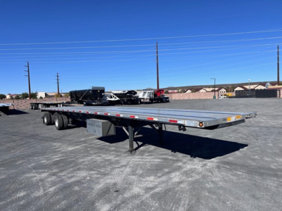 Flatbed trailer in a parking lot showing a long aluminum deck with dual rear axles and support legs.