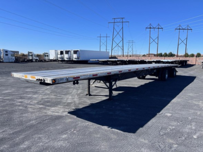 Utility flatbed trailer showing long aluminum deck with a tandem axle, gooseneck hitch not visible, and landing gear, in a lot with power line towers in the background.