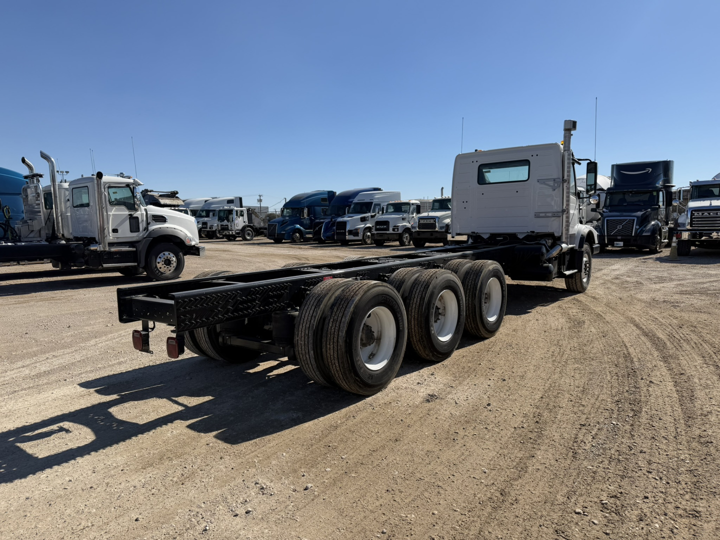 Volvo VHD86B300 cab & chassis truck on a gravel lot, side view showing three rear axles, flatbed frame, and sleeper cab in background, VIN 4V5KH9EH2RN654647.