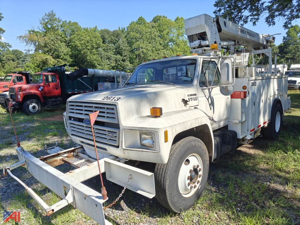 1993 Ford F-700 bucket truck with aerial lift. Front grille, white cab, large bumper with towing arms, bucket crane on rear, service body with compartments. Visible text includes Ford branding on grille and model badges, and equipment label