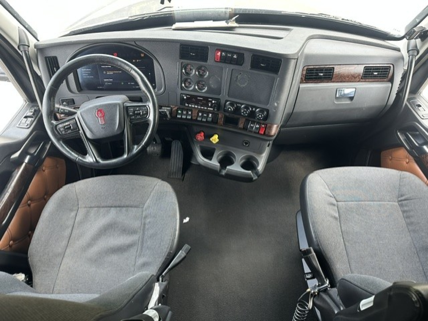 Truck cab interior with steering wheel, dashboard, center console, and two seats; Kenworth logo on steering wheel; multiple gauges and controls visible.