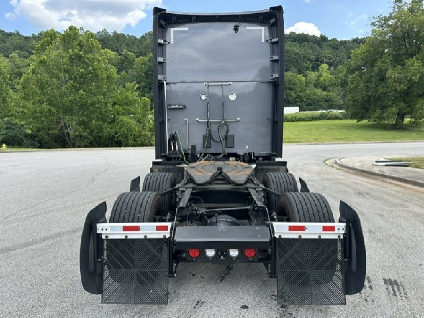 Kenworth T680 rear view showing fifth wheel, dual rear axles, and mud flaps; rear chassis with hoses and electrical connections, gray cabin undercarriage, highway background.