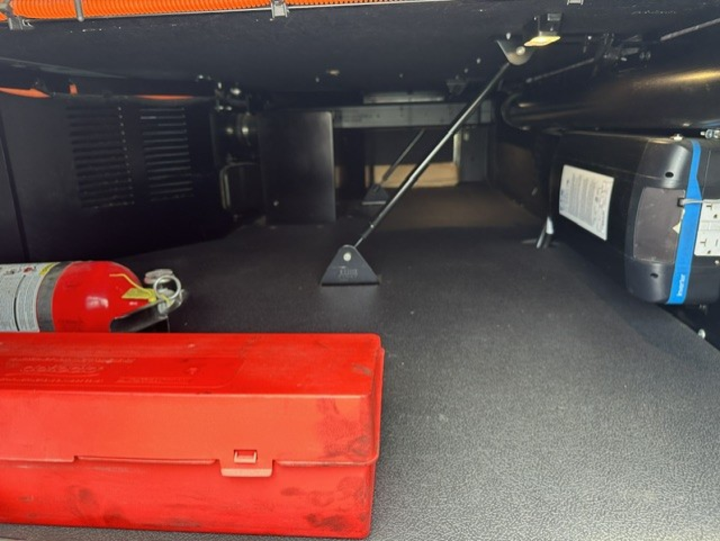 Under-cabinet storage area of a Kenworth T680 showing a red toolbox, a fire extinguisher, a blue OEM battery pack, and a support rod with a triangular foot.
