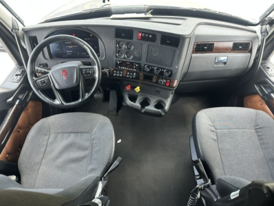 Truck cab interior with steering wheel, dashboard, center console, and two seats; Kenworth logo on steering wheel; multiple gauges and controls visible.