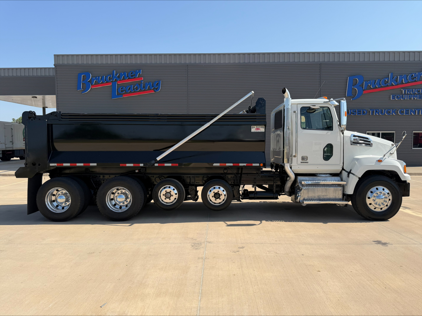 Truck side view of a white 2017 Western Star 4700SF dump truck with black dump bed, parked in front of a building with Bruckner Leasing signage.