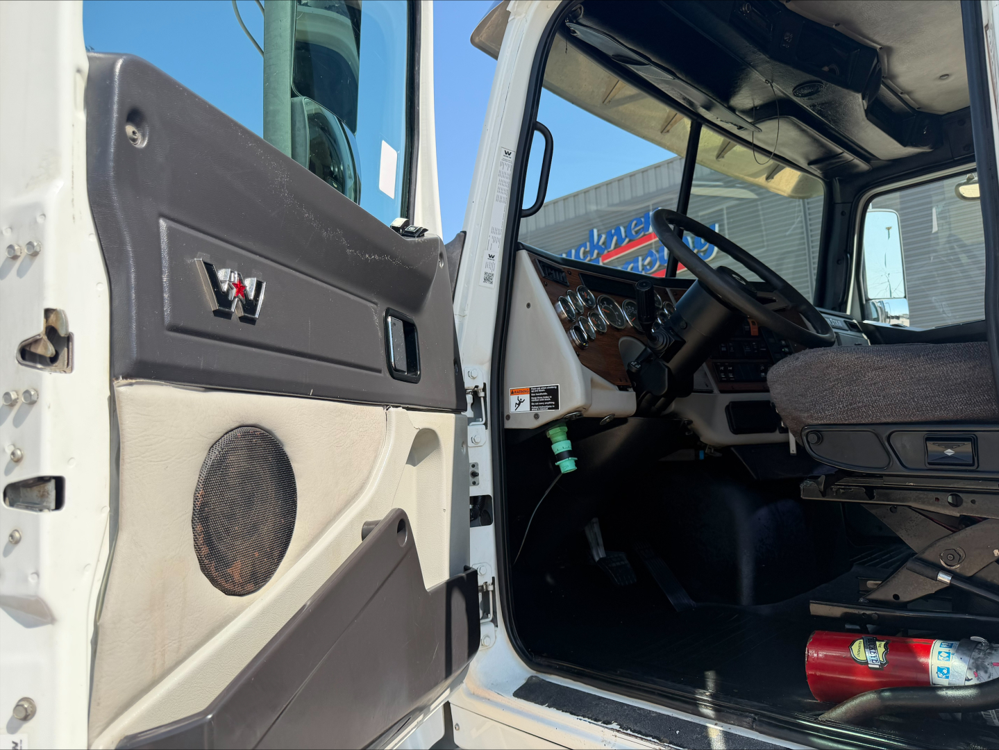 Western Star 4700SF cab interior and open driver door showing dash, steering wheel, and seat with some tools in floor; exterior white door and door panel visible.