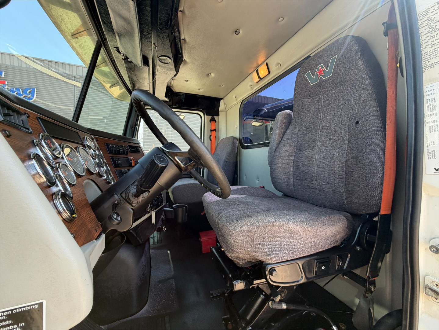 Truck cockpit of a 2017 Western Star 4700SF dump truck showing grey fabric driver seat, steering wheel, dashboard with gauges, and entry door; under-hood area visible.