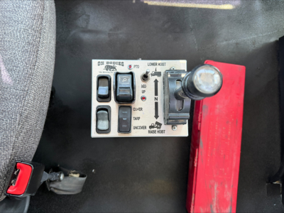 Trucks control panel for dumper with labeled switches and levers; shows 'LOWER HOIST', 'RAISE HOIST', 'CLVER', 'TARP', 'UNCOVER' and 'PTO' with switches and a vertical hoist control lever. Red toolbox to the right.