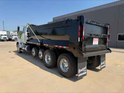 Dump truck in a dealership lot with a black dump body and dual rear axles. Visible text includes warning sticker on tailgate and reflective tape.