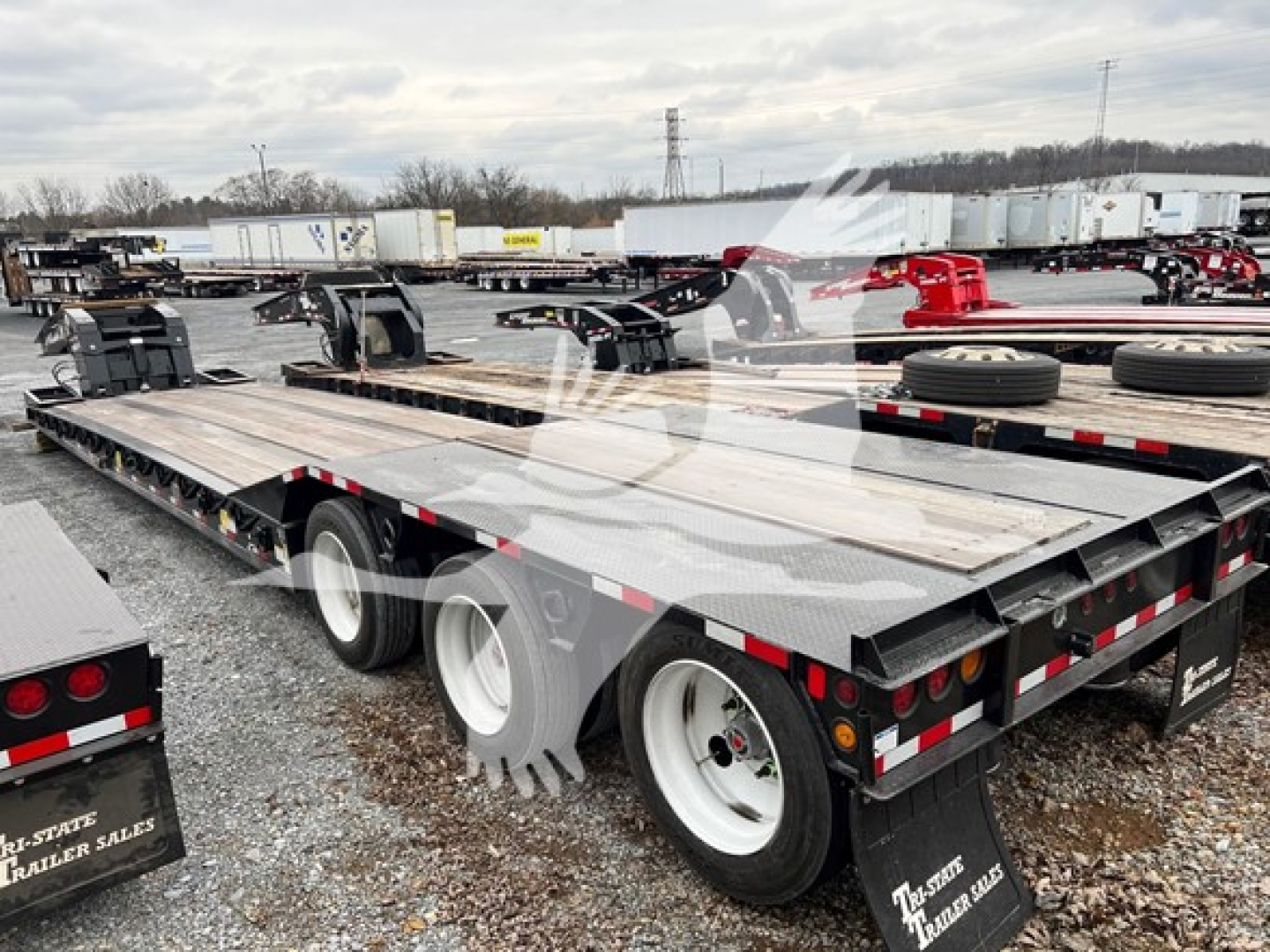 Flatbed trailer showing lowboy deck with multiple axles and tires, chrome wheel rims, and reflective safety tape; background includes other trailers in lot.