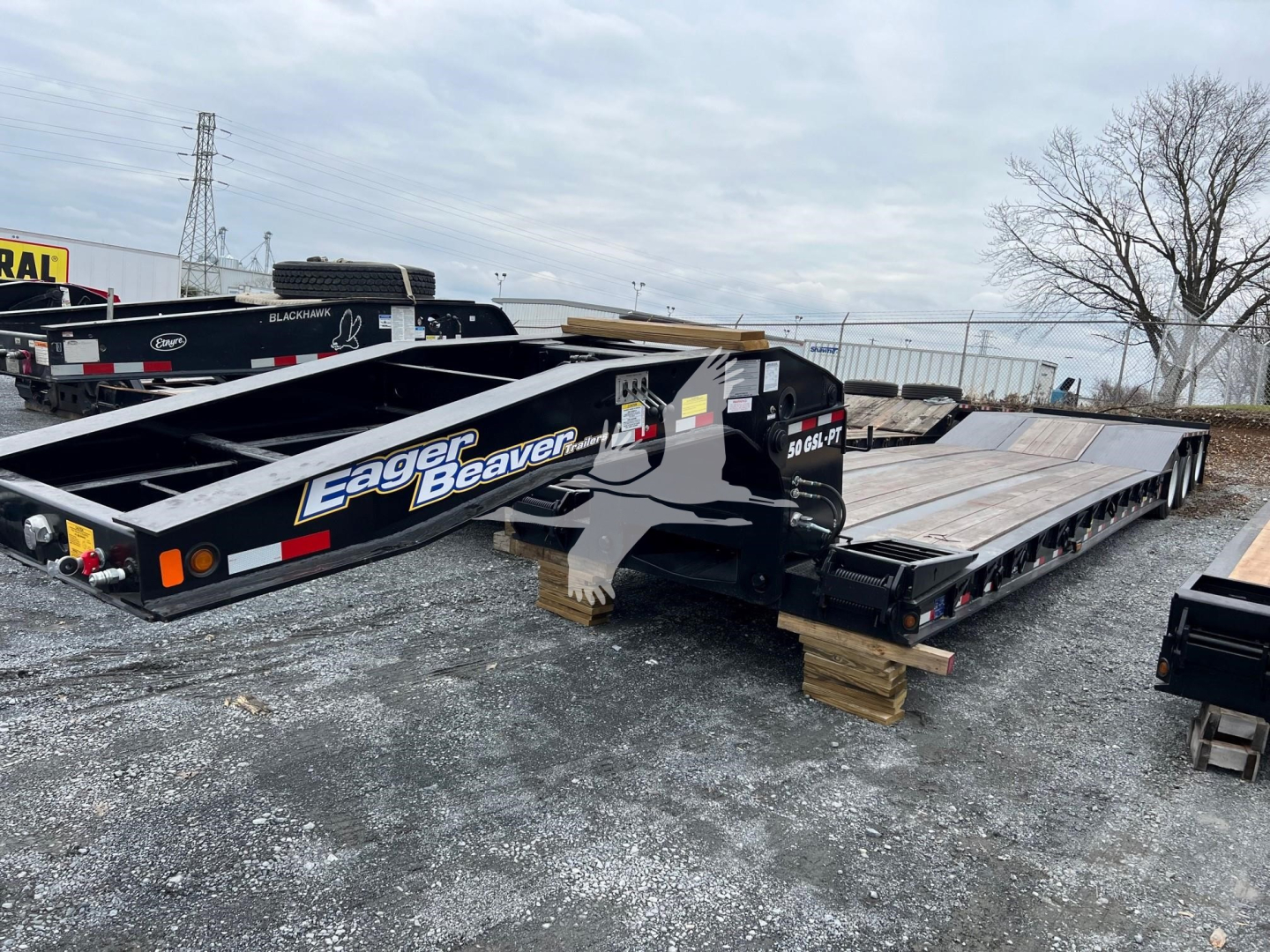 Lowboy trailer showing Eager Beaver branding and twin beams, flat deck with wood blocks under suspension, side guard rails and hydraulic components visible.