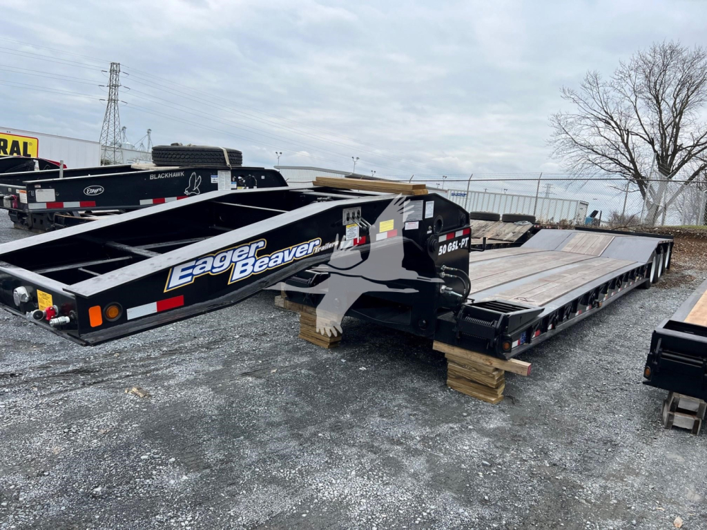 Lowboy trailer showing Eager Beaver branding and twin beams, flat deck with wood blocks under suspension, side guard rails and hydraulic components visible.